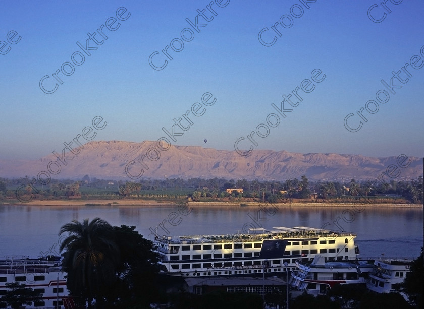 Luxor View EG94538jhp 
 Egypt Luxor Hotel River Nile View balloons dawn West Bank Hatshepsut looking over the quiet River Nile on a sunny September dawn from one of the riverside balconies of this long standing central Luxor Hotel on the city waterfront. This was my first visit to Luxor and I was lucky to get changed to a room in the north wing which was considerably quieter than one in the centre of the hotel above the swimming pool, various restaurants and a dance floor. It also offered great views to the West Bank and in this case two hot air balloons starting their trips over the West Bank. In 1994 the Hotel was the Etap but is now the Mercure and has a great location on the promenade with easy access by foot to the Luxor Museum, to public ferry points for crossing the Nile and to nearby shops or just enjoyed a leisurely walk along the tree lined waterfront by the berthed cruiseboats and feluccas. 
 Keywords: Egypt, Thebes, Luxor, Waset, Ipetisut, East, Bank, River, Nile, Mercure, Etap, Hotel, riverside, riverbank, West, bank, Hatshepsut, temple, balloons, landscape, palm, trees, water, shimmering, September, morning, dawn, sunshine, blue, sky, hazy, Theban, hills, waterfront, promenade, esplanade, modern, architecture, style, stylish, balconies, balcony, tree-lined, papyrus, cruiseboats, berthed, 1994, slide, film, 645, medium, format, transparency, Bronica, ETRSi, scanned, scan