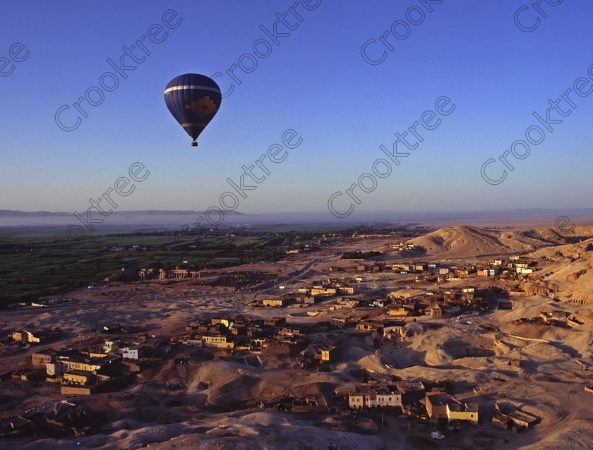 Luxor Balloon EG94217jhp 
 Ramasseum Temple Balloon flight view West bank houses fields tombs hills Egypt the famous temple of Ramesses is located just beneath the balloon and is situated on the West Bank location of the Nile at Luxor The Ramses 11 mortuary temple with its huge engaged Osiris statues of the pharaoh glowing in the late afternoon sunlight and on the right is the fallen seated granite statue of Ramses and a head that supposedly inspired Shelley's poem of Ozymandias with the first pylon in the distance. In this general area are several Tombs of the Nobles which are usually privately organised visits but very well worth the time if you have afew days to spare in Luxor. The balloon flight illustrated here was in 1994 and there were only two although in later years these increased greatly in numbers. This trip took off from the front of Hatshepsut just after dawn, a cold and interesting crossing of the Nile in the dark and long before the modern road bridge and eventually landed in the desert where the Hilton Hotel put on a beautiful breakfast buffet. 
 Keywords: Egypt, Egyptian, Luxor, Ramasseum, Temple, West, Bank, River Nile, Thebes, Qurnat, Murai, Shaykh 'Abd al-Qurnah, landscape, history, archaeology, ancient, Egyptology, hieroglyphics, granite, Osiride, statue, broken, Shelley, Ozymandias, pylon, court, columns, base, hieroglyphs, Ramses, Ramasses, Ramesses, glowing, golden, sunlight, dawn, agriculture, farming, houses, tombs, Nobles, high, panorama, balloon, balloons, high, flying, soaring, 1994, slide, film, 645, medium, format, transparency, scanned, scan, Bronica, ETRSi